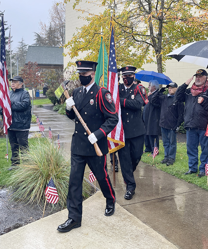 Men in uniform step with flags on a sidewalk.